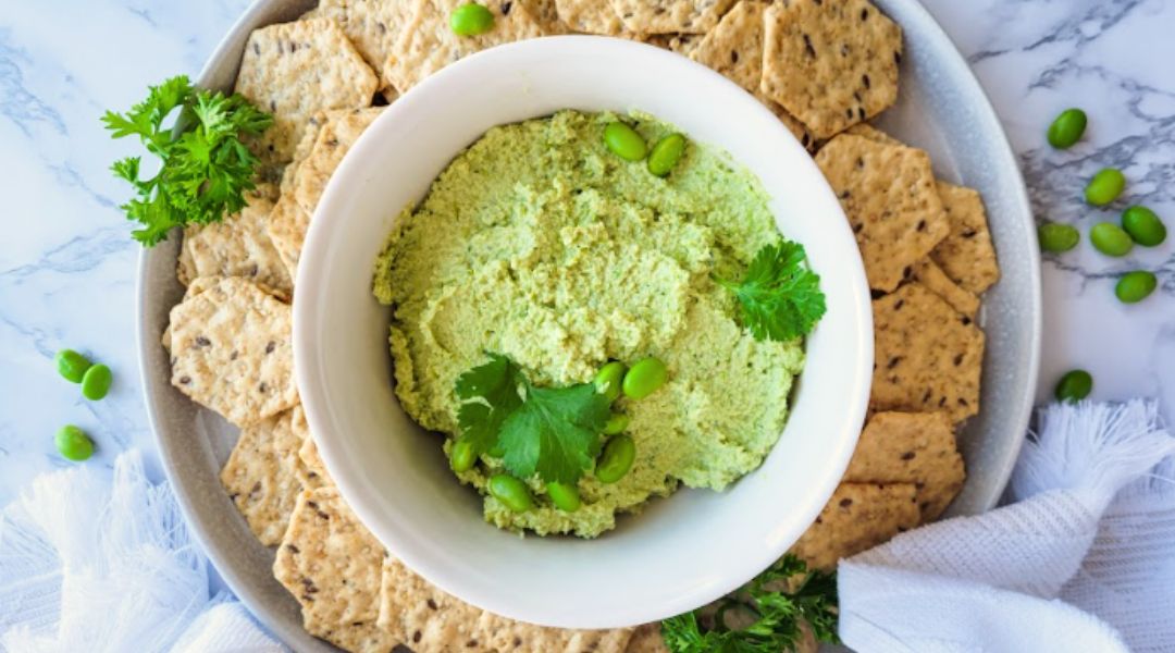 Un bol de houmous d’edamame vert garni de persil, entouré de craquelins aux graines