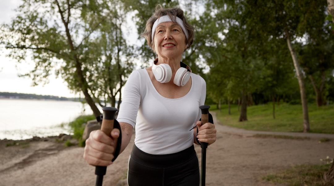 Une femme âgée marche avec des bâtons de marche nordique dans un cadre naturel, affichant un sourire confiant et équipé d’écouteurs.