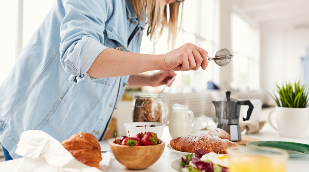 Une personne en chemise en jean bleu clair saupoudre du sucre glace sur un croissant à une table de petit-déjeuner garnie de pâtisseries, de fruits et de café.