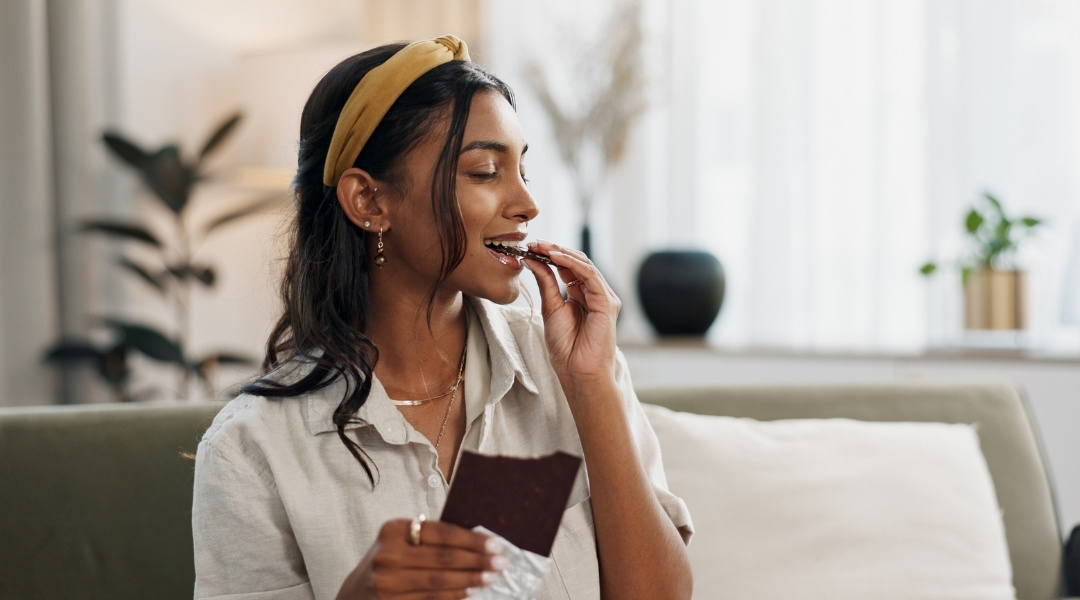 Une femme souriante avec un bandeau moutarde savoure un morceau de chocolat noir assise sur un canapé dans un salon confortable.