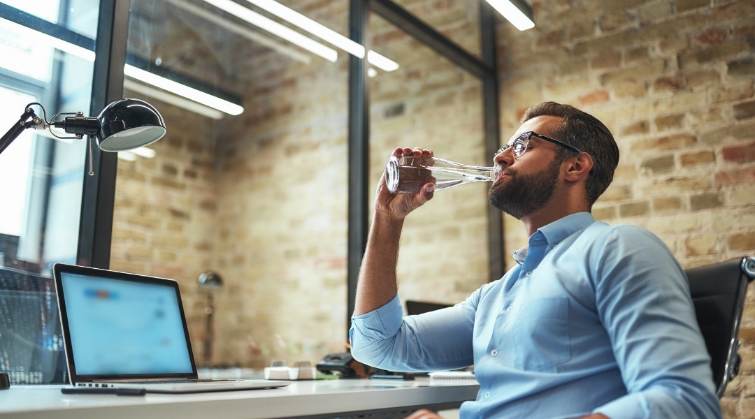 Un homme en chemise bleue et lunettes boit de l'eau dans une bouteille en verre assis à un bureau dans un bureau moderne.