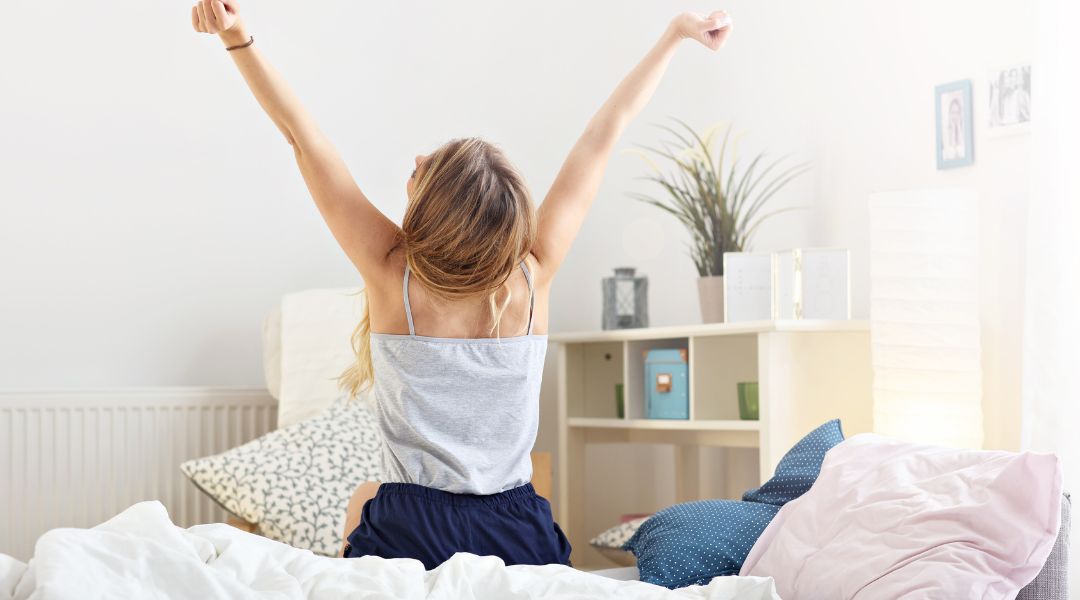 Une femme s'étirant les bras après s'être réveillée, assise sur un lit dans une chambre lumineuse et minimaliste avec lumière naturelle.