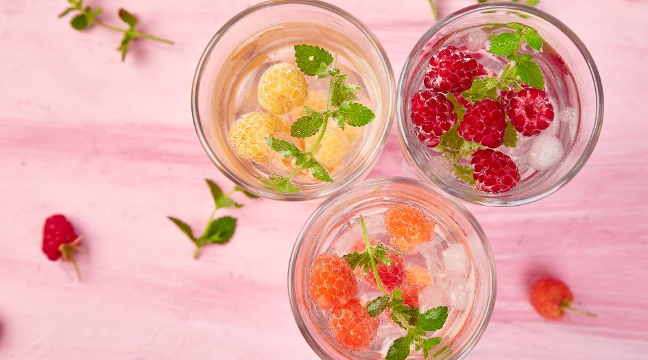 Trois verres d'eau infusée avec des framboises jaunes, rouges et orange, des feuilles de menthe et des glaçons, sur un fond rose.