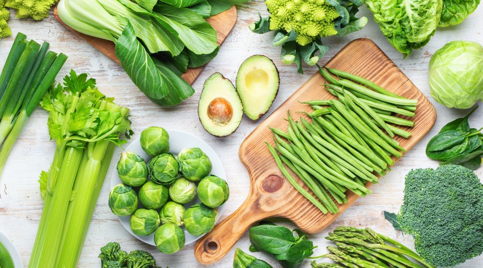 Un assortiment de légumes verts frais, dont du céleri, des haricots verts, de l'avocat et de la laitue, disposés sur une surface en bois clair.