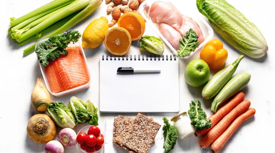 Un carnet vierge entouré d'aliments sains tels que du saumon, du poulet, des fruits, des légumes et des noix, disposés sur un fond blanc.