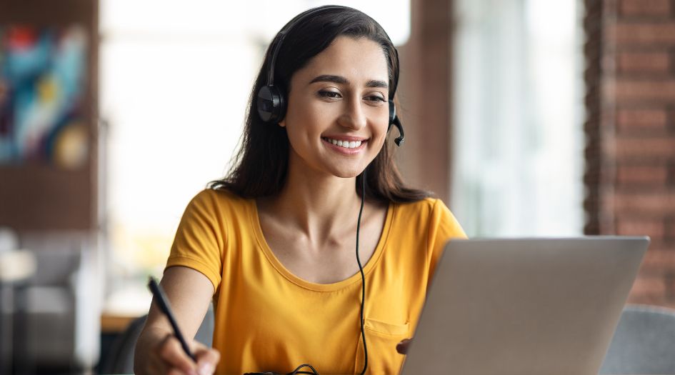 Une femme souriante en chemise jaune, portant un casque et utilisant un ordinateur portable, assise dans un intérieur lumineux.