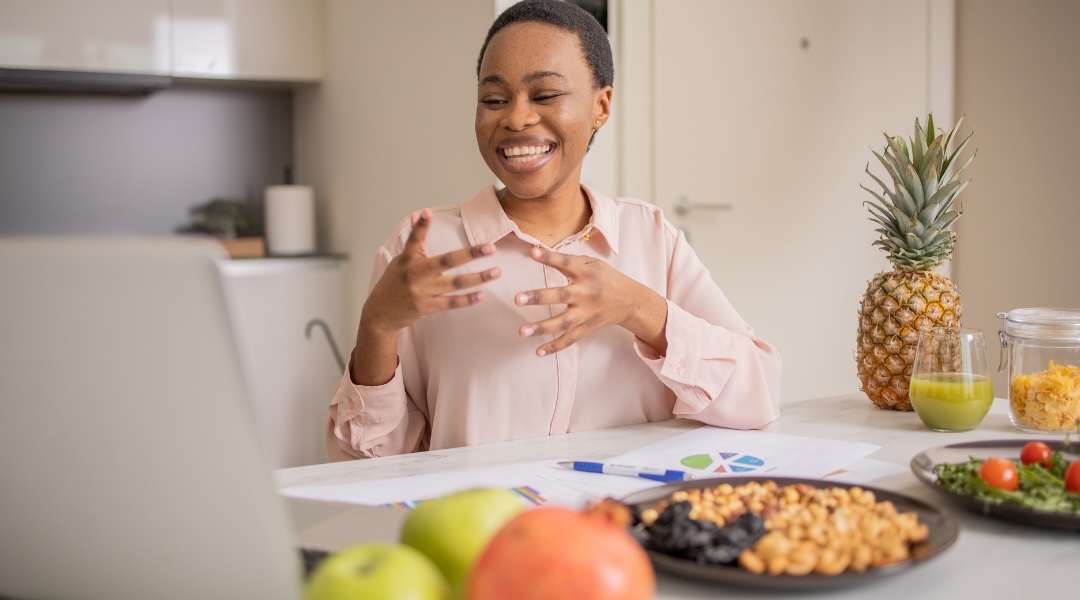 Une femme souriante participe à une consultation en nutrition en ligne, entourée d’aliments sains et de documents de travail.