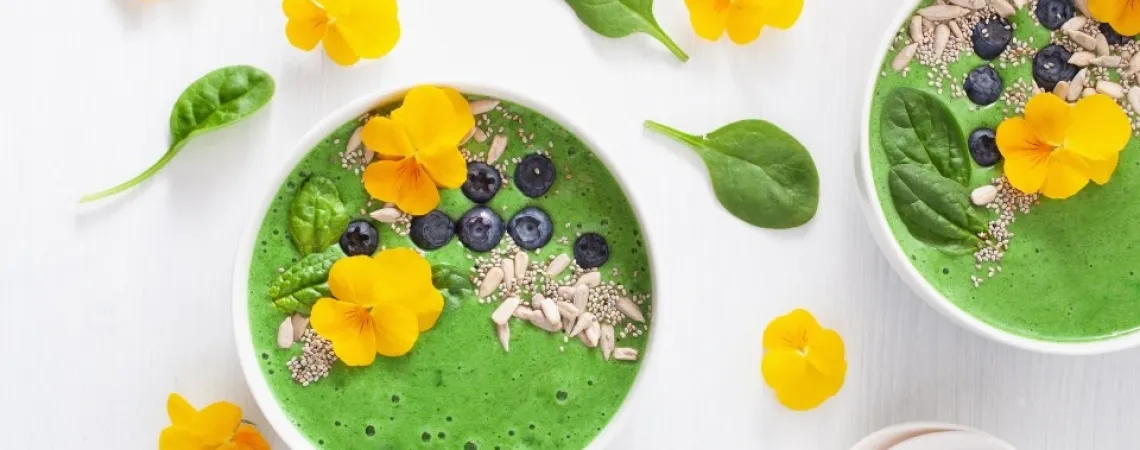 Un bol de smoothie vert vif garni de fleurs comestibles jaunes, de myrtilles fraîches, de graines de chia, de graines de tournesol et de feuilles de jeunes épinards. Le bol est disposé sur un fond clair avec des feuilles d'épinards et des fleurs jaunes supplémentaires éparpillées en décoration.