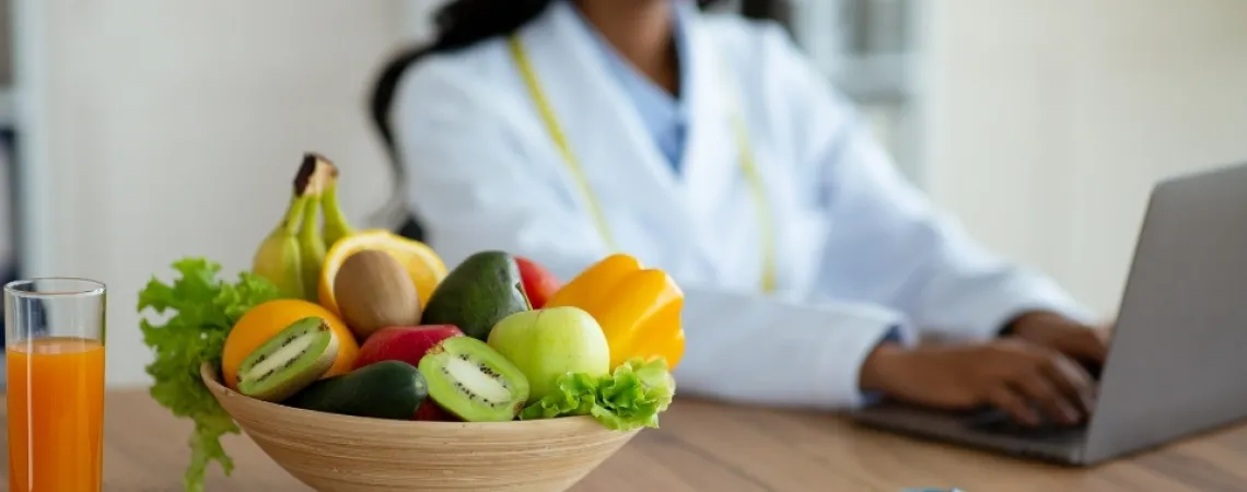 Un bol de fruits frais, dont kiwi, pomme et banane, avec un verre de jus sur une table, tandis qu'une nutritionniste travaille sur un ordinateur portable en arrière-plan.