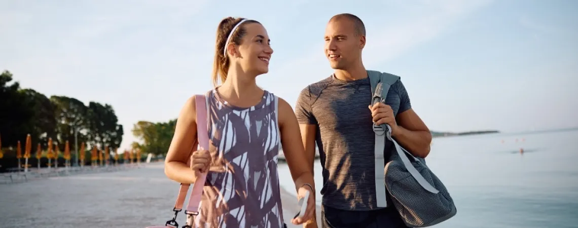 Un couple souriant en tenue de sport marche sur une plage, chacun portant un sac de sport, avec la mer et des arbres en arrière-plan.