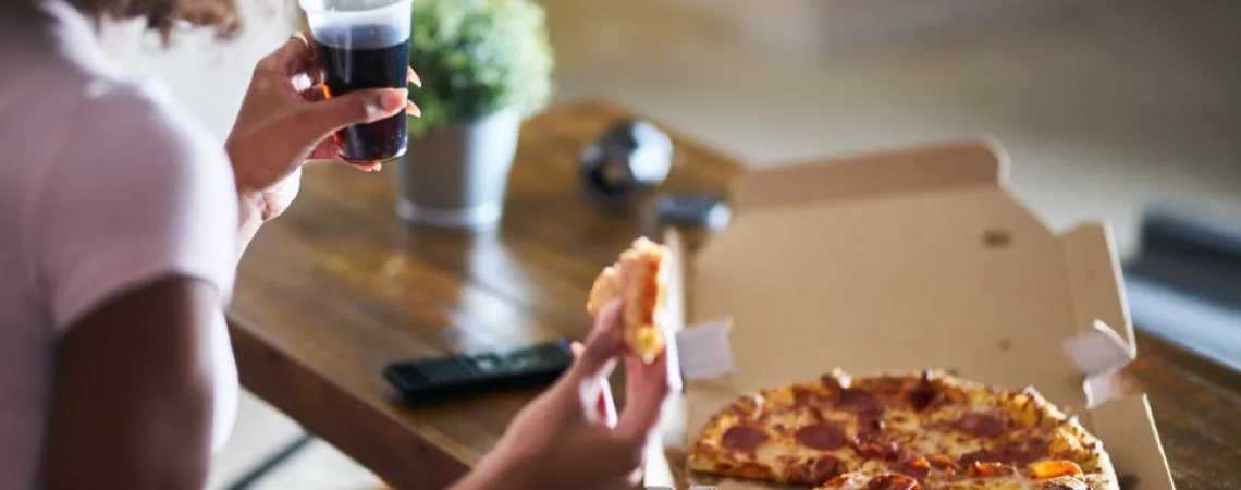 Personne tenant une part de pizza au pepperoni et un verre de soda, avec une boîte à pizza sur une table en bois.