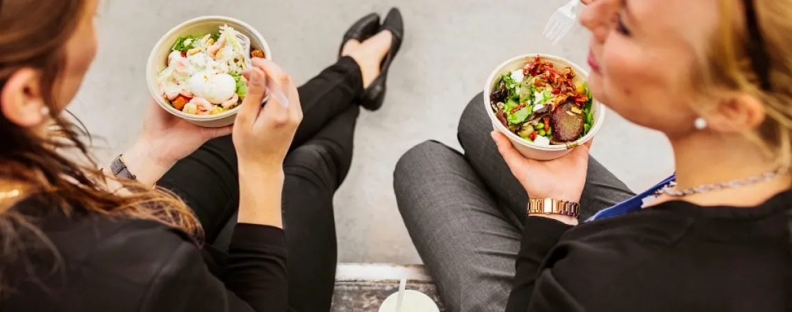 Deux femmes assises à l'extérieur, mangeant des bols de salade fraîche avec des légumes verts et garnitures, accompagnées de boissons.