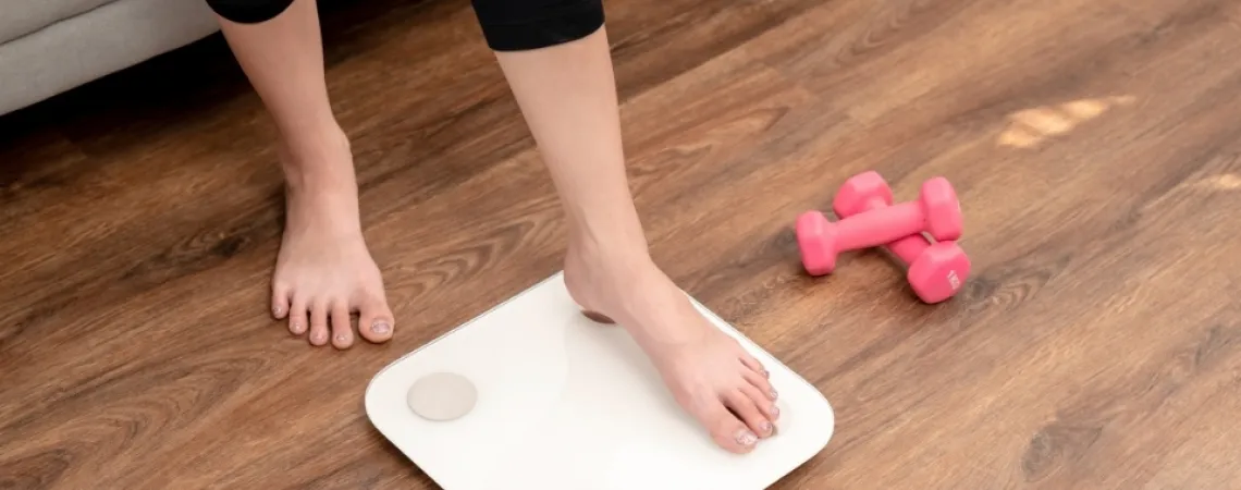 Personne montant sur une balance blanche dans un salon avec des haltères roses sur le sol en bois à proximité.