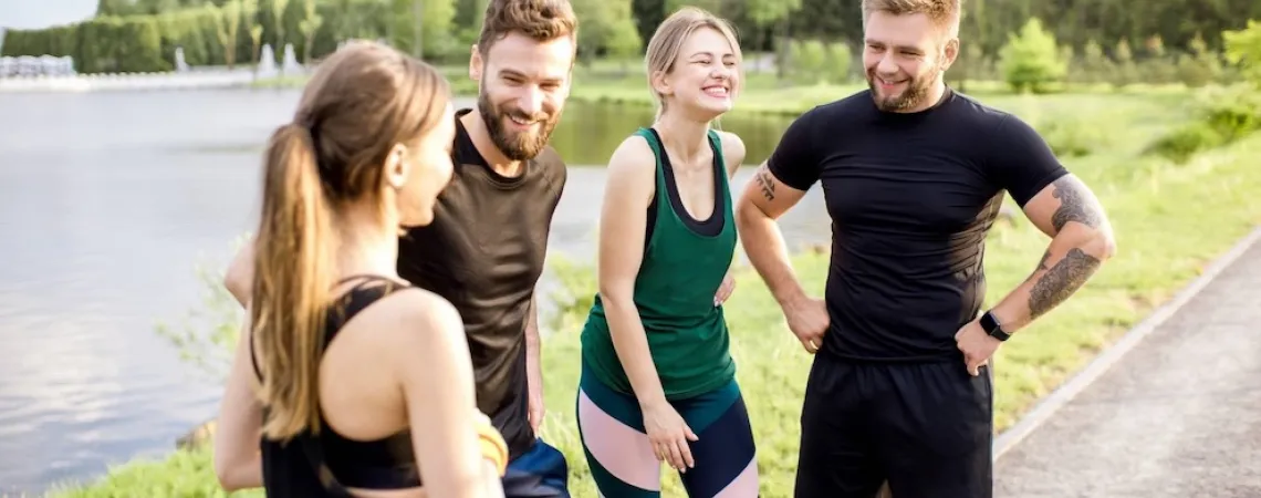Un groupe de quatre amis en tenue de sport souriant et discutant à l'extérieur près d'un lac, avec une zone boisée en arrière-plan.