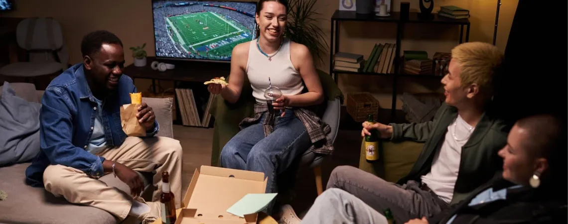 Quatre amis rient et mangent de la pizza dans un salon confortable en regardant un match de football américain à la télévision