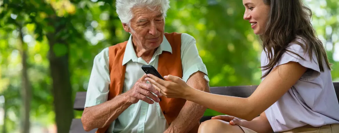 A young woman showing an elderly man how to use a smartphone while they sit on a park bench. The man has a continuous glucose monitor on his arm.