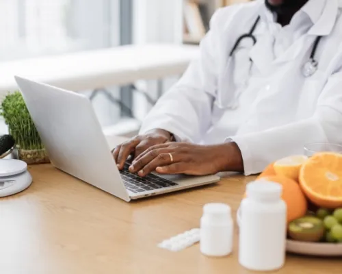 Un nutritionniste diététiste en blouse blanche travaille sur un ordinateur portable avec des fruits, légumes et suppléments sur la table.