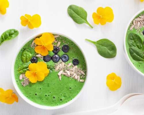 Un bol de smoothie vert vif garni de fleurs comestibles jaunes, de myrtilles fraîches, de graines de chia, de graines de tournesol et de feuilles de jeunes épinards. Le bol est disposé sur un fond clair avec des feuilles d'épinards et des fleurs jaunes supplémentaires éparpillées en décoration.