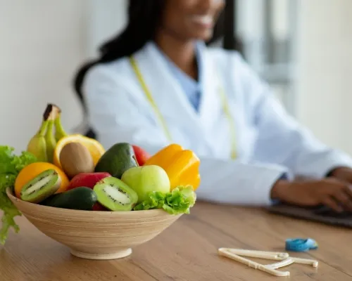 Un bol de fruits frais, dont kiwi, pomme et banane, avec un verre de jus sur une table, tandis qu'une nutritionniste travaille sur un ordinateur portable en arrière-plan.