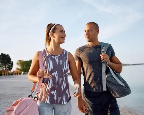 Un couple souriant en tenue de sport marche sur une plage, chacun portant un sac de sport, avec la mer et des arbres en arrière-plan.