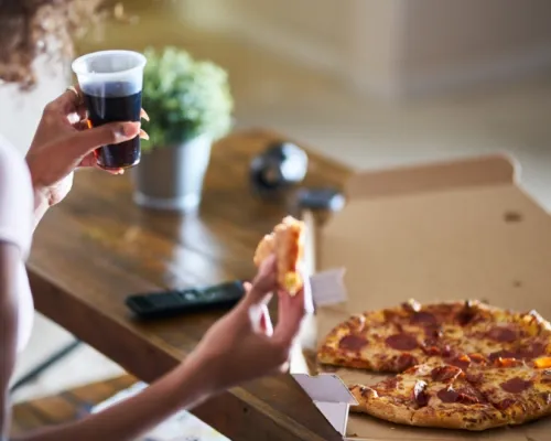 Personne tenant une part de pizza au pepperoni et un verre de soda, avec une boîte à pizza sur une table en bois.