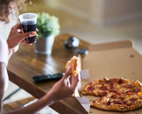 Personne tenant une part de pizza au pepperoni et un verre de soda, avec une boîte à pizza sur une table en bois.