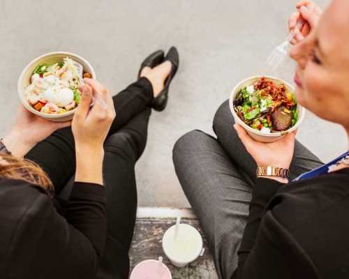 Deux femmes assises à l'extérieur, mangeant des bols de salade fraîche avec des légumes verts et garnitures, accompagnées de boissons.