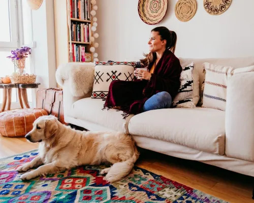Femme assise sur un canapé avec une tasse, un chien couché sur un tapis coloré devant elle - Woman sitting on a couch with a cup, a dog lying on a colorful rug in front of her