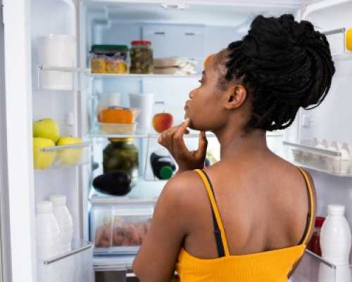 Femme debout devant un réfrigérateur ouvert, réfléchissant en regardant différents aliments et boissons à l'intérieur.  Woman standing in front of an open fridge, thoughtfully looking inside at various food items and drinks.