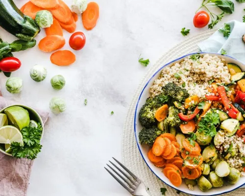 Colorful chopped vegetables scattered on a light background, with a bowl of couscous topped with broccoli, zucchini, carrots, tomatoes, chickpeas, and herbs.