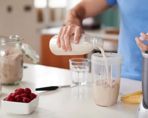 A person in a blue shirt pours a protein shake into a blender cup - Une personne en t-shirt bleu verse un shake de protéines dans un gobelet