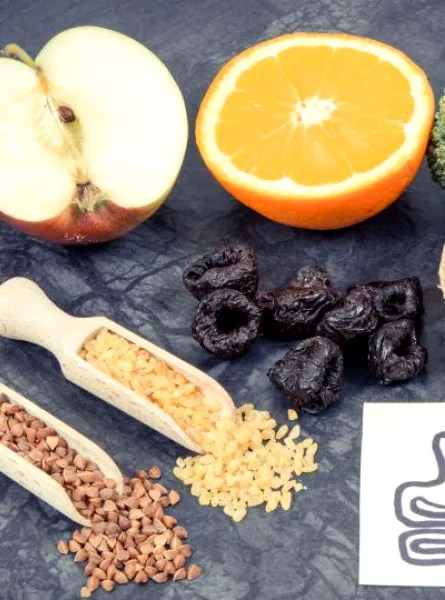Aliments riches en fibres incluant des fruits comme la pomme et l'orange, des légumes comme le brocoli, du pain complet, des abricots secs, des pruneaux, ainsi que des céréales telles que le sarrasin et le riz brun, posés à côté d'une illustration d'un estomac pour représenter les possibles inconforts liés à la présence de FODMAP