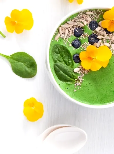 Un bol de smoothie vert vif garni de fleurs comestibles jaunes, de myrtilles fraîches, de graines de chia, de graines de tournesol et de feuilles de jeunes épinards. Le bol est disposé sur un fond clair avec des feuilles d'épinards et des fleurs jaunes supplémentaires éparpillées en décoration.