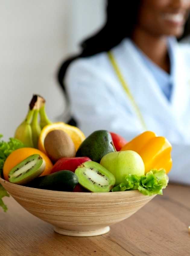 Un bol de fruits frais, dont kiwi, pomme et banane, avec un verre de jus sur une table, tandis qu'une nutritionniste travaille sur un ordinateur portable en arrière-plan.