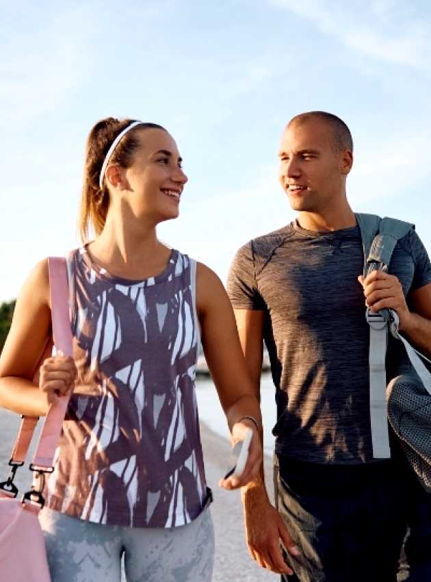 Un couple souriant en tenue de sport marche sur une plage, chacun portant un sac de sport, avec la mer et des arbres en arrière-plan.