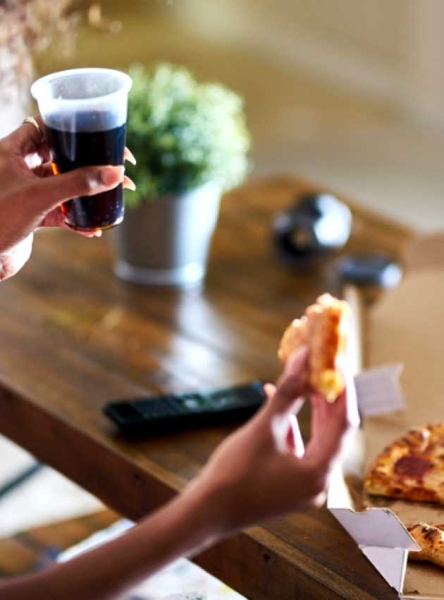 Personne tenant une part de pizza au pepperoni et un verre de soda, avec une boîte à pizza sur une table en bois.