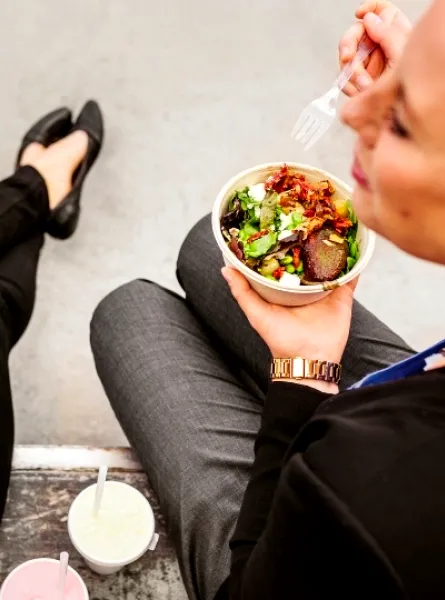 Deux femmes assises à l'extérieur, mangeant des bols de salade fraîche avec des légumes verts et garnitures, accompagnées de boissons.