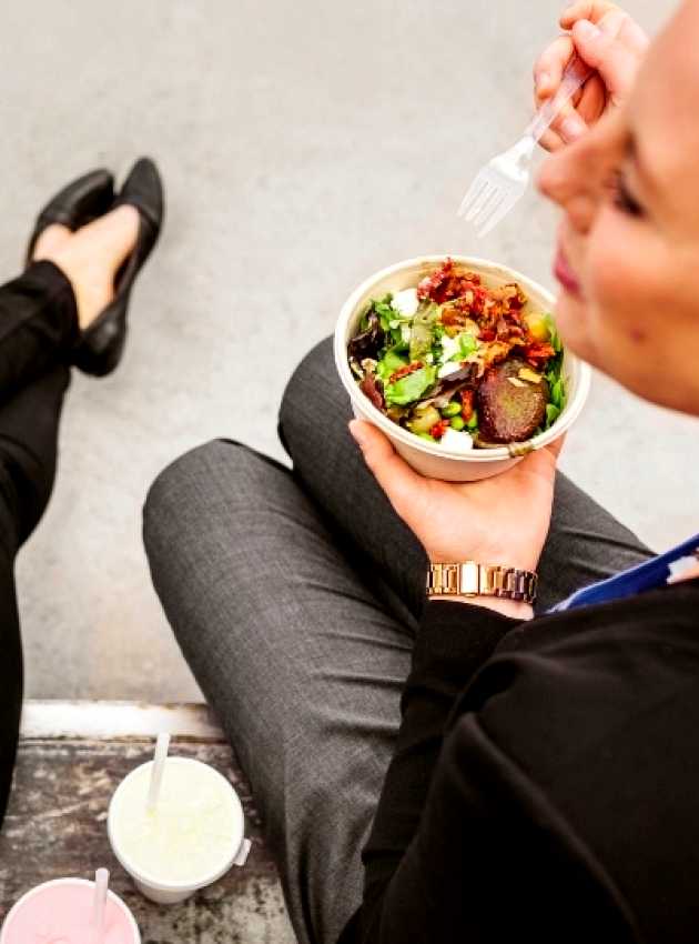 Deux femmes assises à l'extérieur, mangeant des bols de salade fraîche avec des légumes verts et garnitures, accompagnées de boissons.