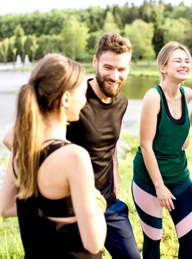 Un groupe de quatre amis en tenue de sport souriant et discutant à l'extérieur près d'un lac, avec une zone boisée en arrière-plan.