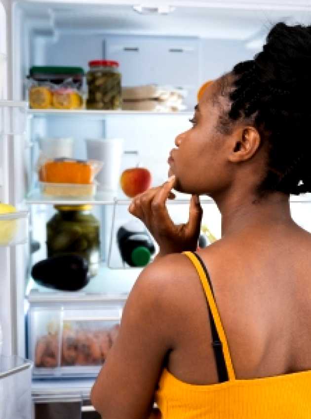 Femme debout devant un réfrigérateur ouvert, réfléchissant en regardant différents aliments et boissons à l'intérieur.  Woman standing in front of an open fridge, thoughtfully looking inside at various food items and drinks.