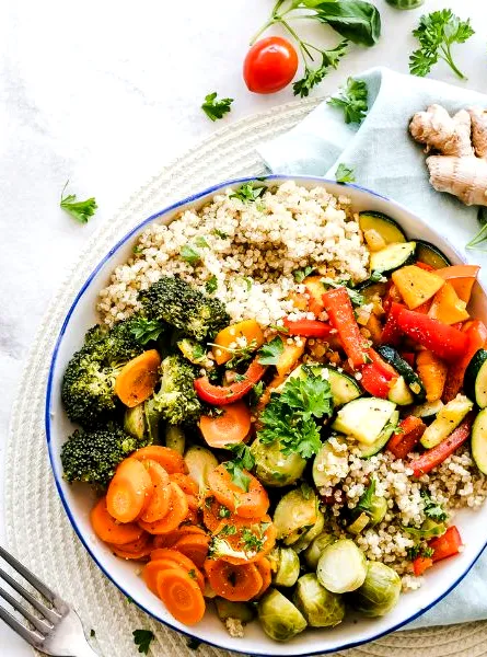 Colorful chopped vegetables scattered on a light background, with a bowl of couscous topped with broccoli, zucchini, carrots, tomatoes, chickpeas, and herbs.