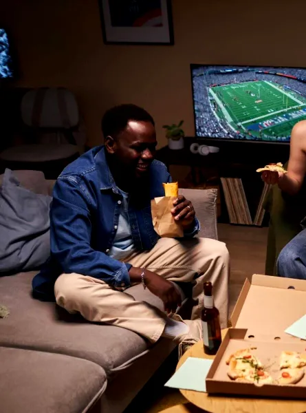 Quatre amis rient et mangent de la pizza dans un salon confortable en regardant un match de football américain à la télévision