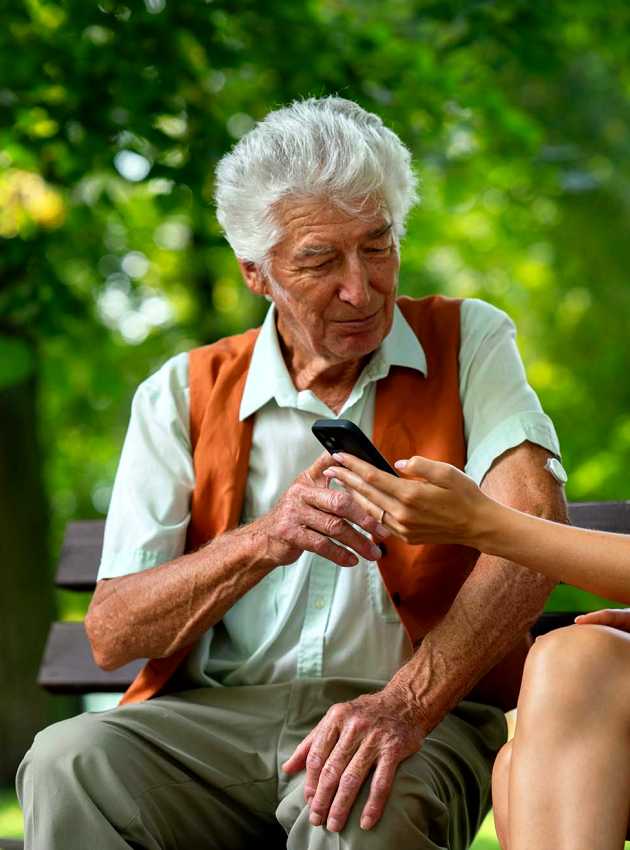 A young woman showing an elderly man how to use a smartphone while they sit on a park bench. The man has a continuous glucose monitor on his arm.